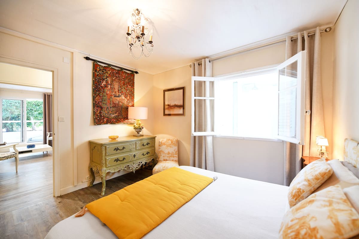 A bedroom with a bed, a nightstand, a lamp, a window with curtains, a chair, a dresser, and a tapestry on the wall.