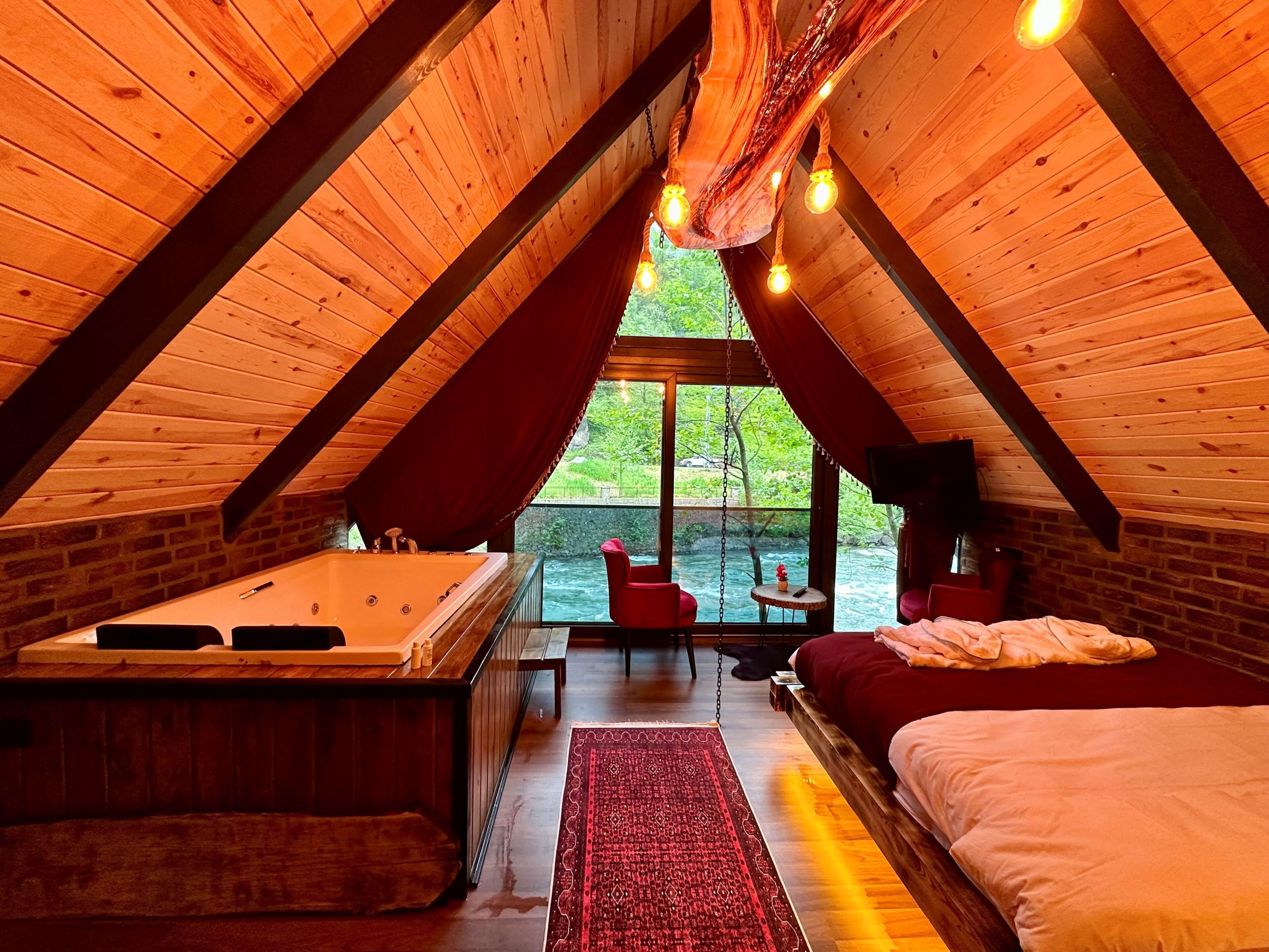 An attic bedroom with a wooden sloped ceiling, a hot tub, a bed, and a view of greenery outside.