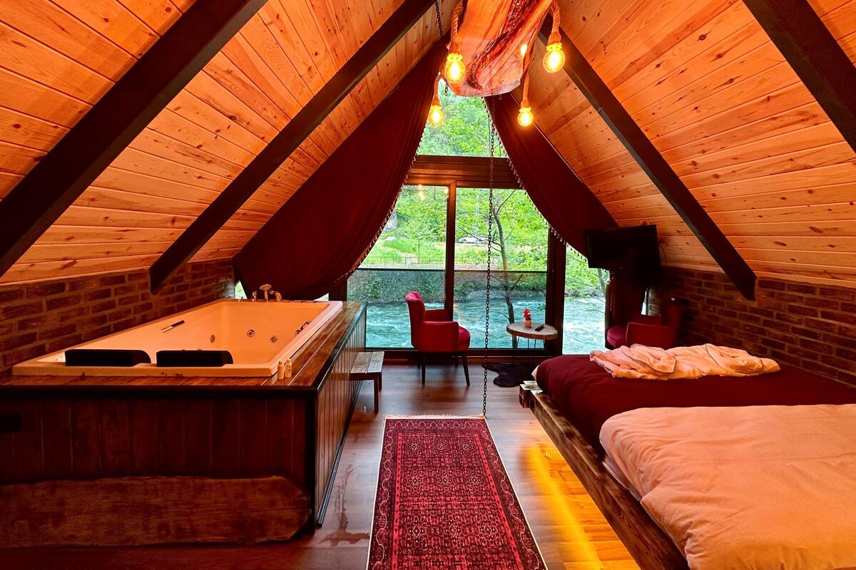 An attic bedroom with a wooden sloped ceiling, a hot tub, a bed, and a view of greenery outside.