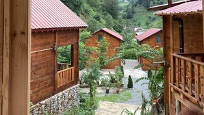 A wooden cabin with a stone foundation, surrounded by lush greenery and a mountainous backdrop. - Tanura Bungalows (Camlihemsin)