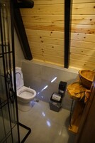 Un baño pequeño y funcional con pared de madera, inodoro blanco, un cubo de basura y lavamanos de madera.