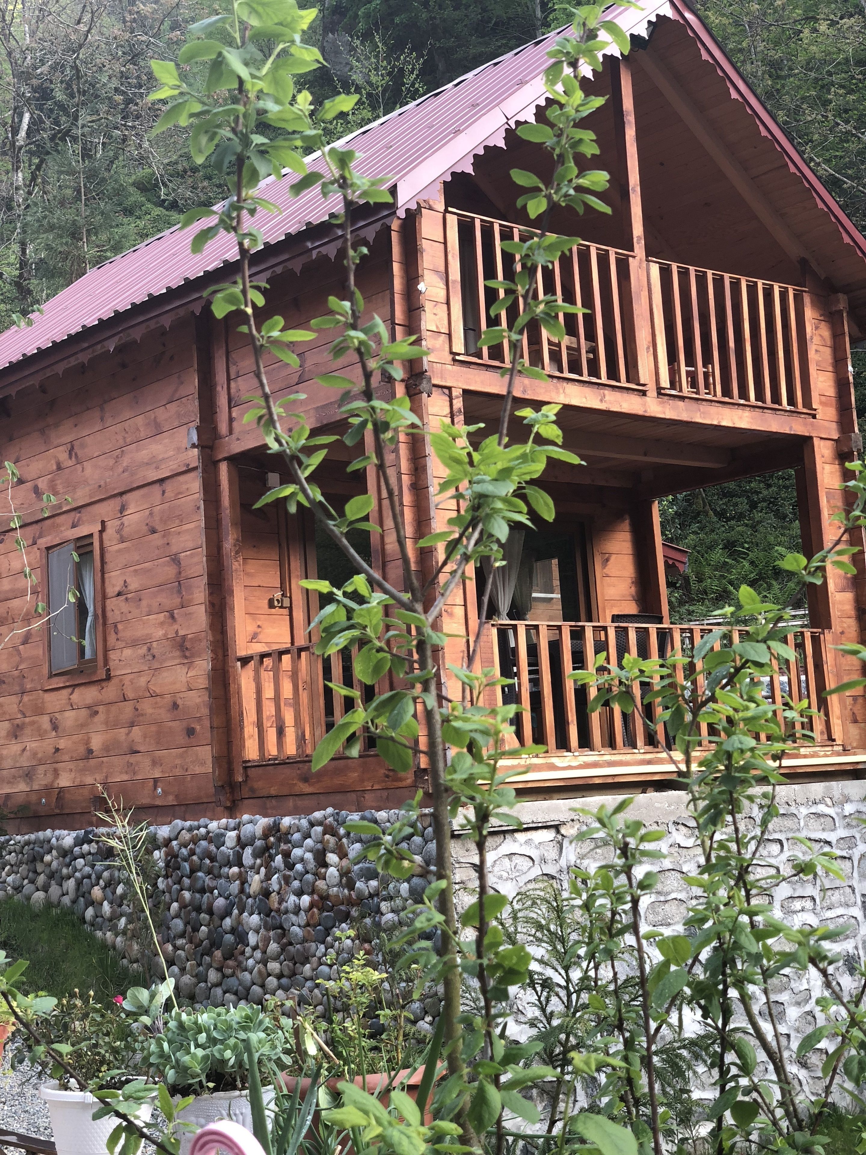 Une cabane en bois dotée d’un balcon, entourée de verdure et bordée d’un mur de pierres.