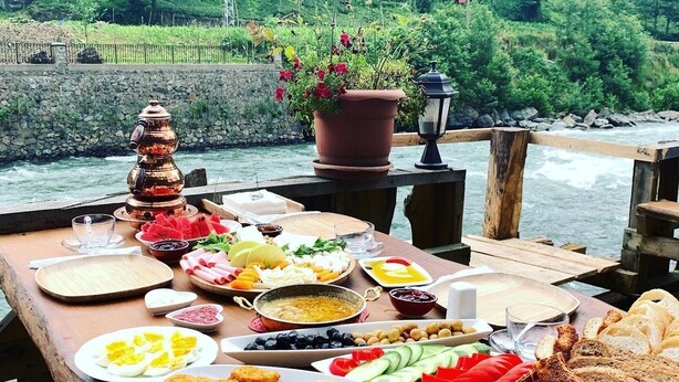 A picnic table with various food items and a potted plant, set against a backdrop of a river and greenery. - Tanura Bungalows (Camlihemsin)