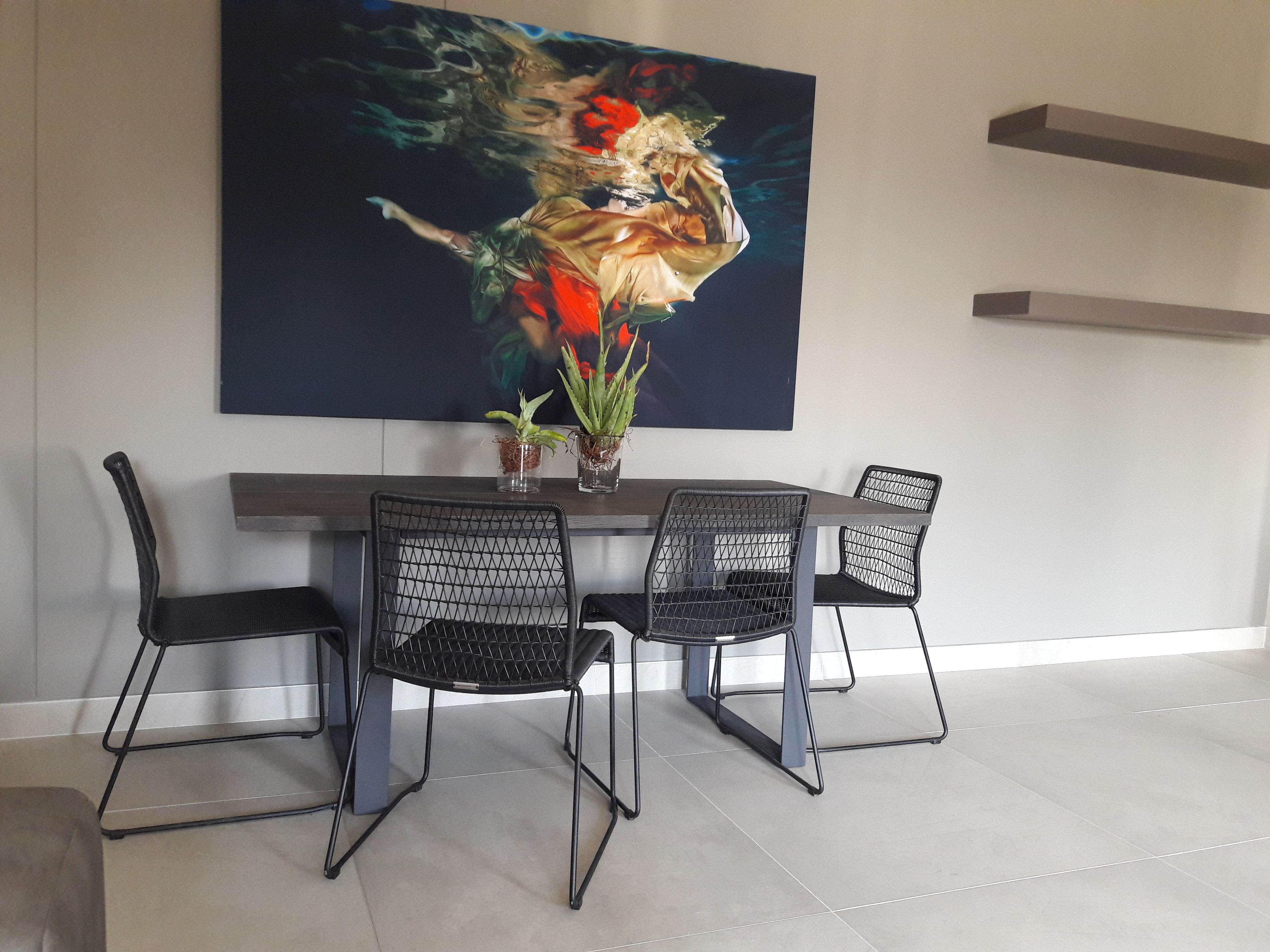Un coin repas avec une table moderne et quatre chaises noires à assise en treillis. Un grand tableau abstrait est accroché au mur, au-dessus de la table.