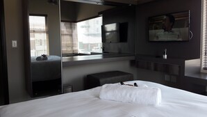 A modern hotel room with a large bed, a built-in bench, a mirror, and a TV. - Atlantic Apartments (Swakopmund)
