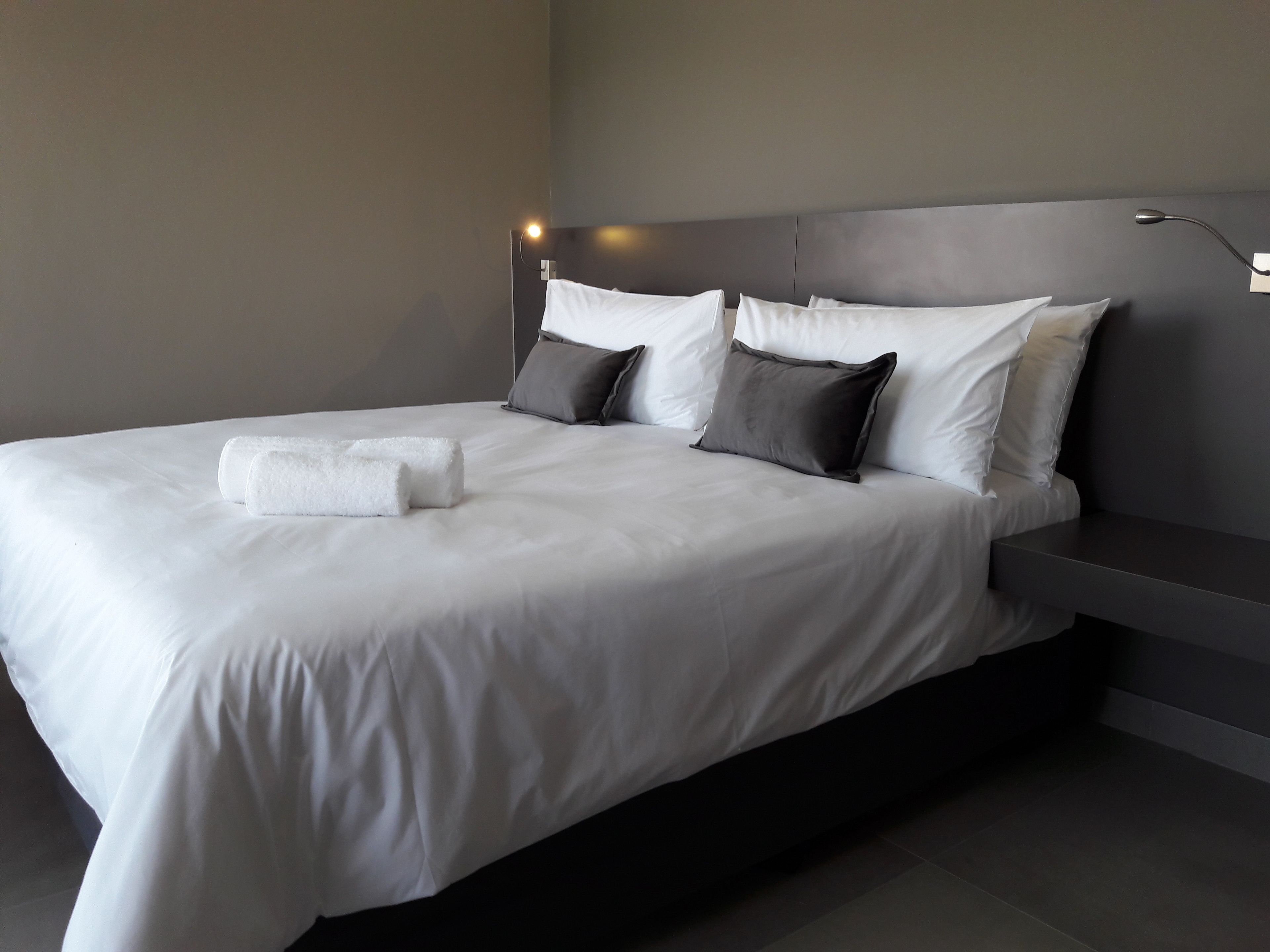 Quarto de hotel moderno com uma cama grande, cabeceira cinza e uma mesinha de cabeceira.