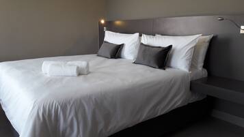 Quarto de hotel moderno com uma cama grande, cabeceira cinza e uma mesinha de cabeceira.