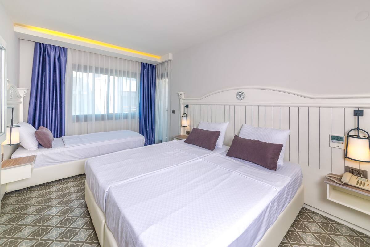 A hotel room with two beds, a large window with blue curtains, a bedside table with a lamp, and a wall-mounted thermostat.