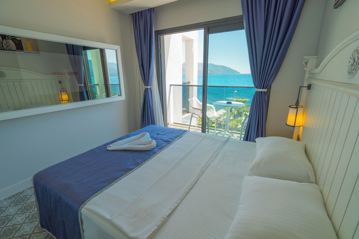 Una habitación de hotel con cama, un balcón con vistas al mar y una ventana con cortinas.