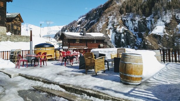 A wooden building with a snow-covered roof, outdoor seating with red chairs, and wooden barrels. - Pension du Lac Bleu (Evolène)