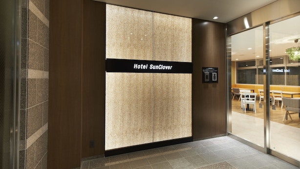 L’entrée d’un hôtel au design moderne, avec une grande enseigne indiquant « Hôtel SunClover », une porte en verre donnant sur un espace intérieur et un sol carrelé.