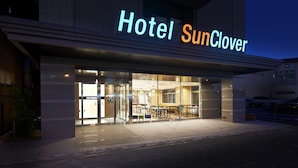 L’entrée d’un hôtel, avec une enseigne indiquant « Hôtel SunClover », des portes vitrées et un espace aménagé avec des sièges à l’extérieur.