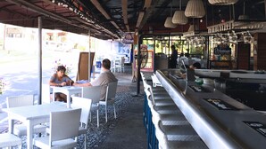 An outdoor cafe with wicker tables and chairs, a bar area with a stainless steel counter, and a view of the street in the background.