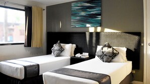 A hotel room with two beds, a large window with curtains, a wall-mounted artwork, and a bedside table with a lamp.