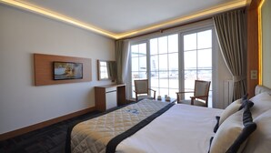 Habitación de hotel con una cama grande, un escritorio, sillas y vistas a la ciudad a través de amplias ventanas.