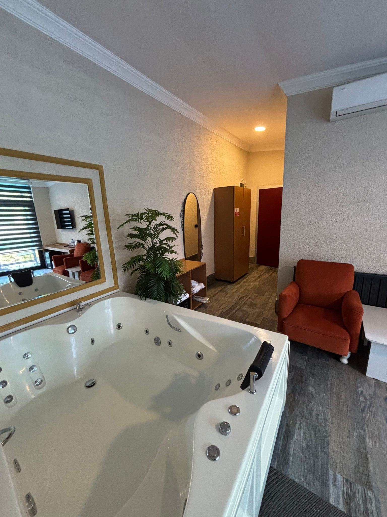 Habitación de hotel con una bañera blanca grande, un sillón rojo, una planta en maceta y un espejo que refleja la habitación.