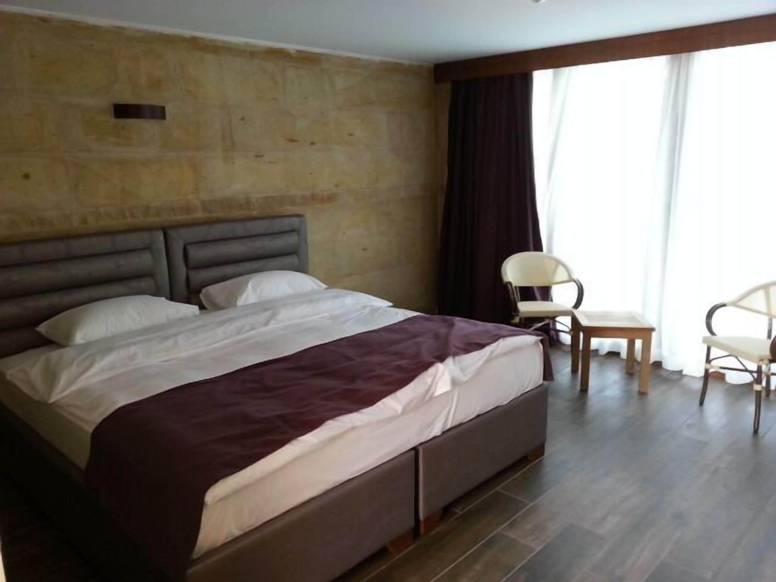 Ein Schlafzimmer mit einem Bett, zwei Stühlen, Holzboden und einem großen Fenster mit Vorhängen.