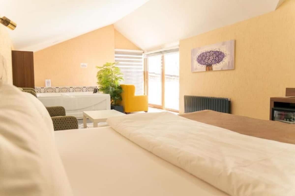 A hotel room with a bed, a small table, a yellow chair, a plant, and a painting on the wall.
