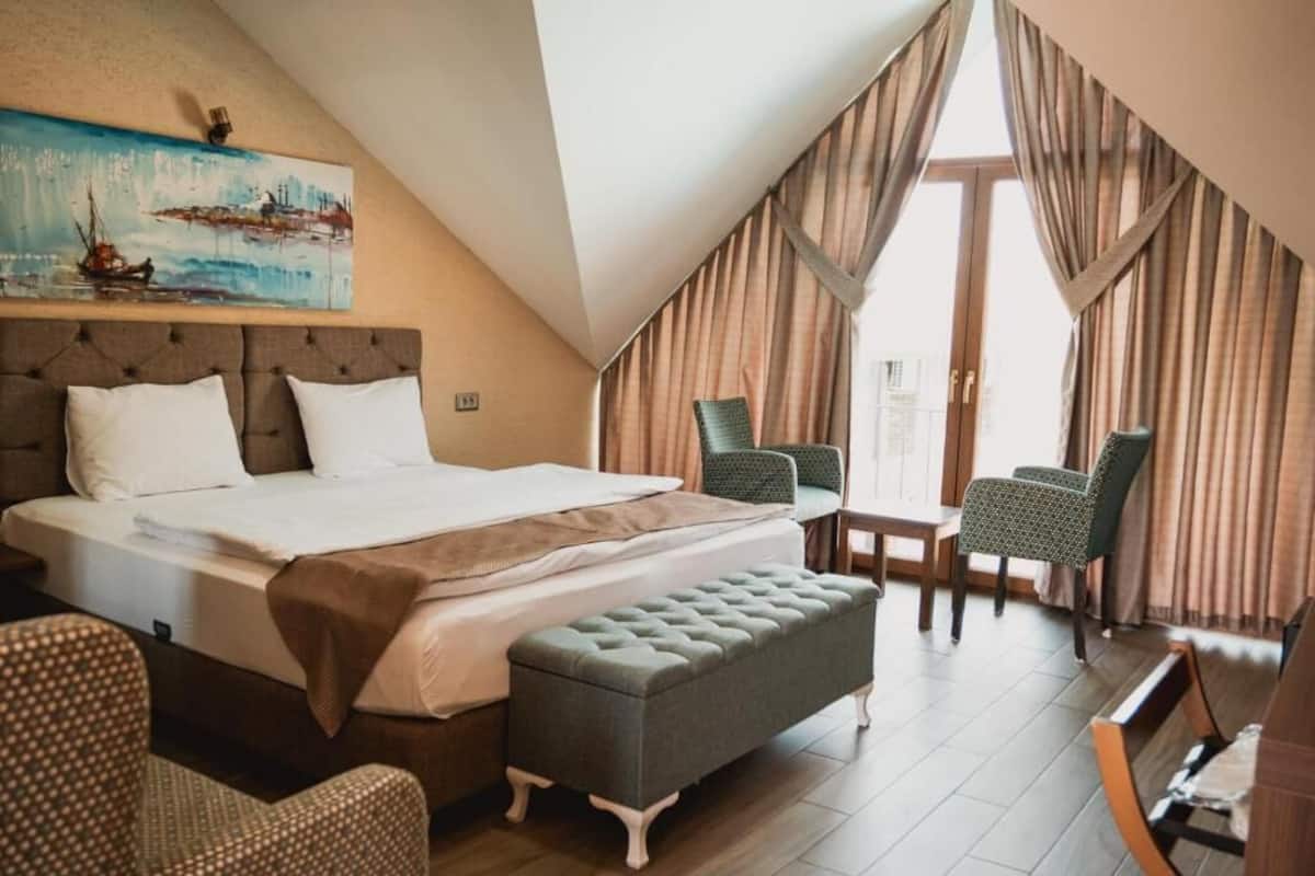 A hotel room with a large bed, a bench, two chairs, a table, and a painting on the wall.