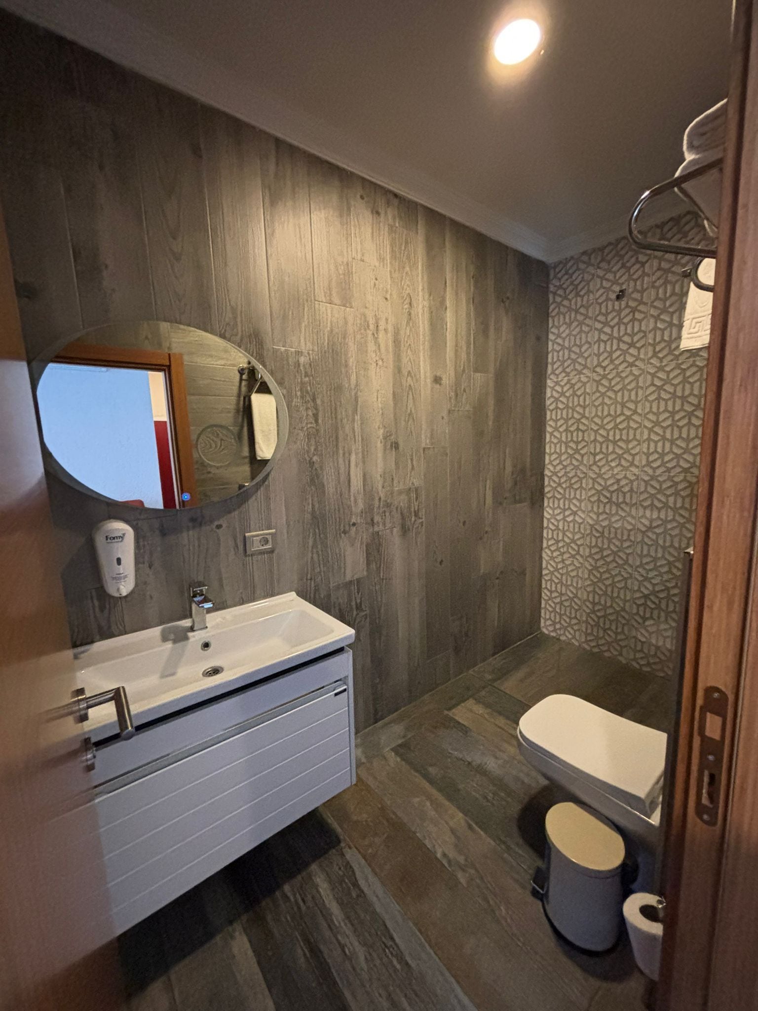 A modern bathroom with a wooden wall, a round mirror, a white sink, and a toilet.
