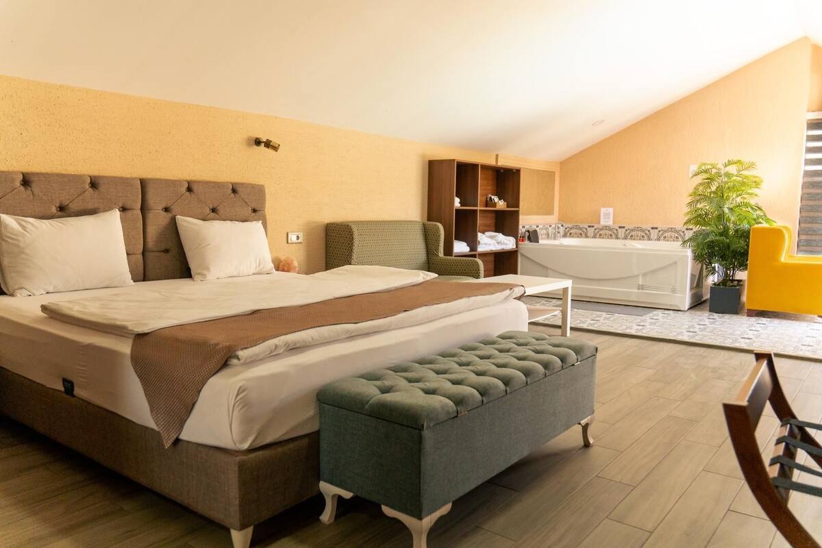 A bedroom with a bed, a bench, a chair, a small table, a shelf, a plant, and a bathtub.