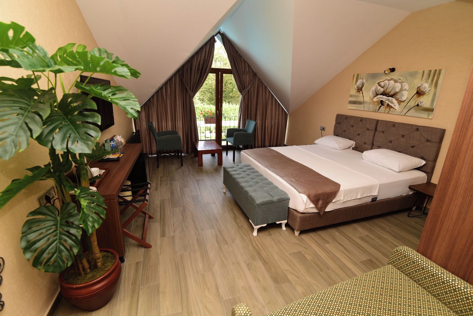 A bedroom with a large bed, a desk, a chair, a potted plant, and a view of the outdoors.
