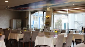 A dining area with tables set for four, a large window with a view of buildings, and a ceiling mural. - Colonia Park Hotel (Sebinkarahisar)