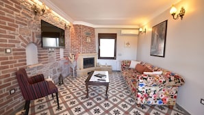 A living room with a brick wall, a fireplace, a sofa, a coffee table, and a television mounted on the wall. - Cunda Rooms (Ayvalik)