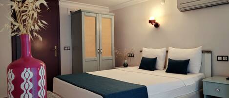 A bedroom with a bed, bedside table, nightstand, wall-mounted air conditioner, and a decorative vase with dried plants.