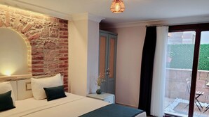 A bedroom with a brick wall, a bed with white bedding and green pillows, a nightstand with a vase, and a sliding glass door leading to an outdoor area. - Cunda Rooms (Ayvalik)