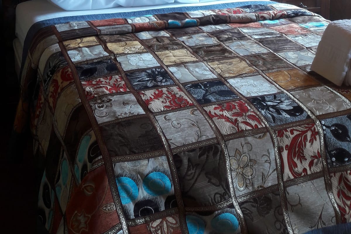Ein Schlafzimmer mit Steinwand, Holzboden und einem Bett mit Patchworkdecke. Es gibt außerdem ein kleines Regal mit einer Statue.
