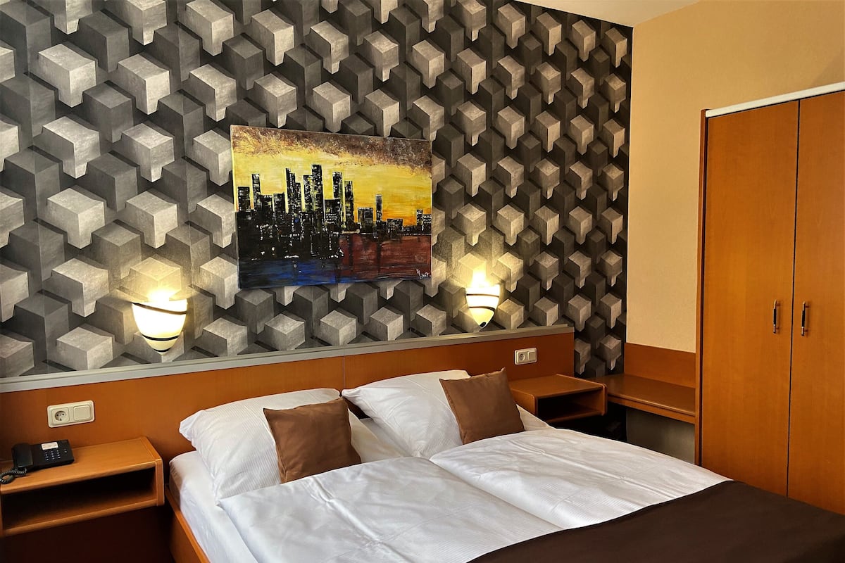 Une chambre d’hôtel avec un lit, des tables de chevet, un tableau représentant un paysage urbain et une armoire.