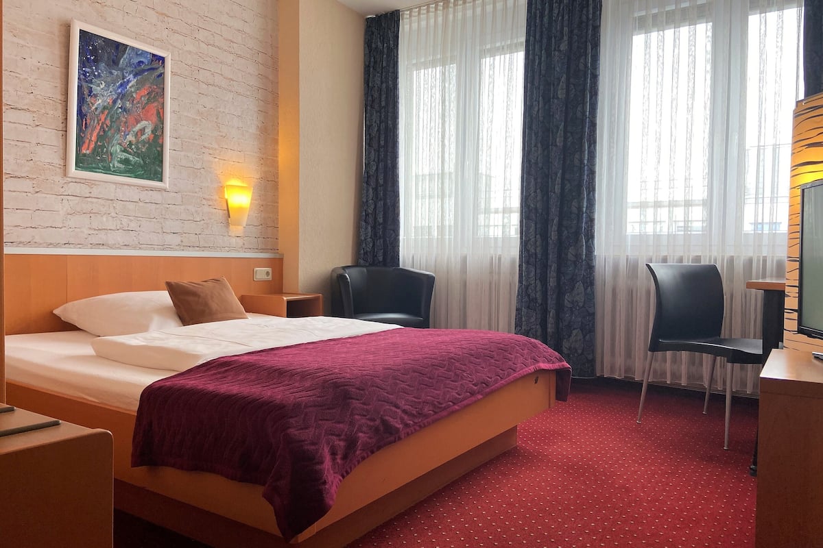 Une chambre d’hôtel avec un lit, un bureau, une chaise, une télévision, un tableau au mur et une fenêtre avec des rideaux.
