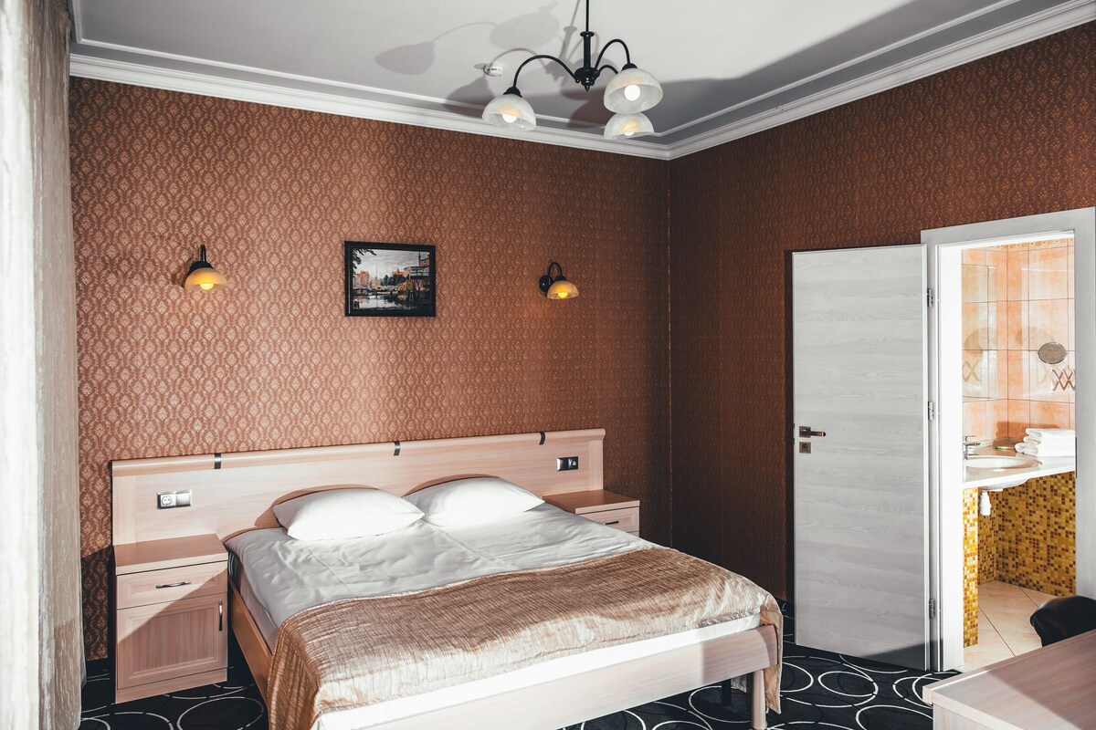 Habitación de hotel con cama, mesitas de noche, una lámpara de pared y una puerta que da al baño.