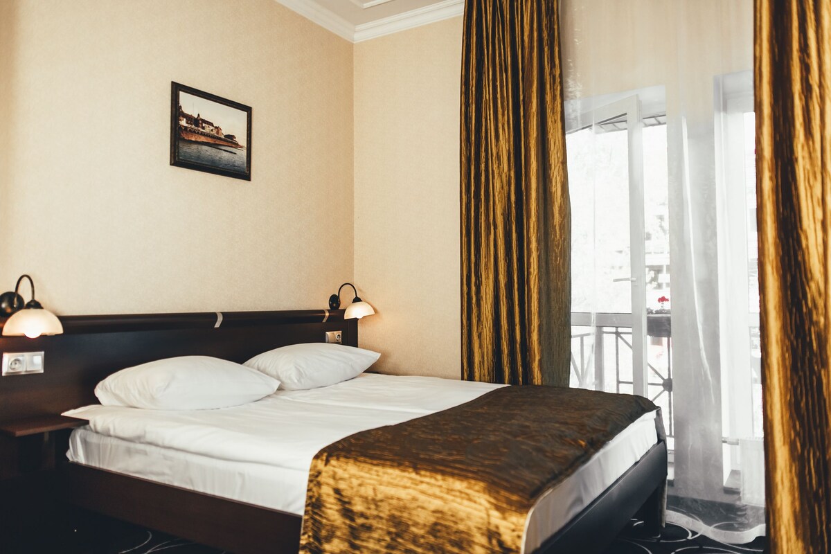 Habitación de hotel con cama, dos lámparas de noche, un cuadro en la pared y vistas a un balcón con cortinas.