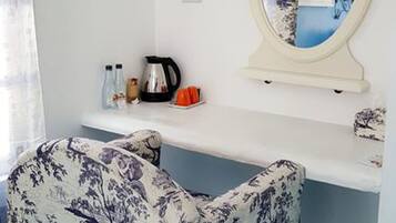 Una habitación con un escritorio blanco, una silla con estampado floral, un espejo, una tetera y una cafetera.