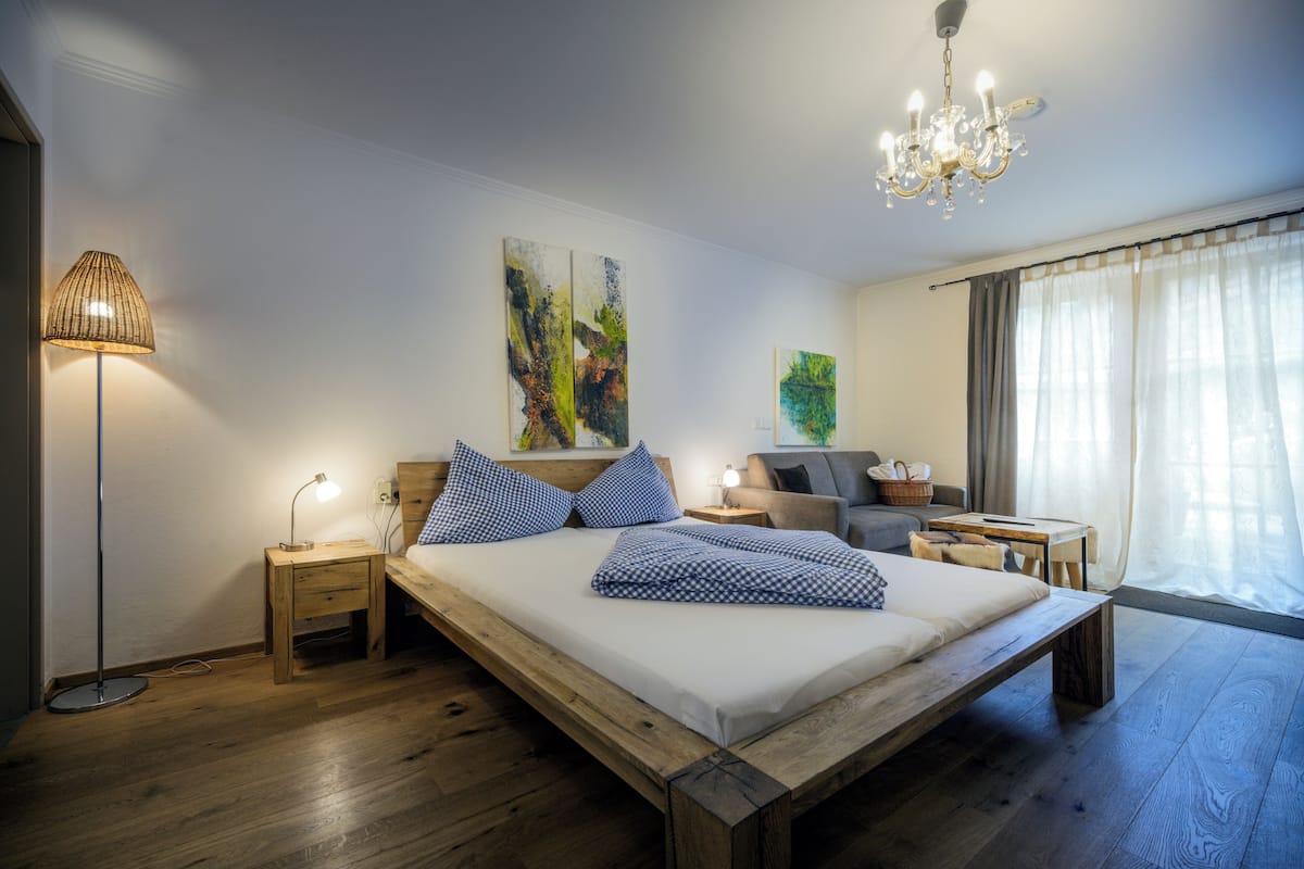 A bedroom with a wooden bed, bedside tables, a lamp, artwork, a sofa, and a window with curtains.