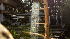 A wooden water feature with water cascading down, surrounded by a garden and a building with a balcony. - Flora - Das kleine Gartenhotel (Ellmau)