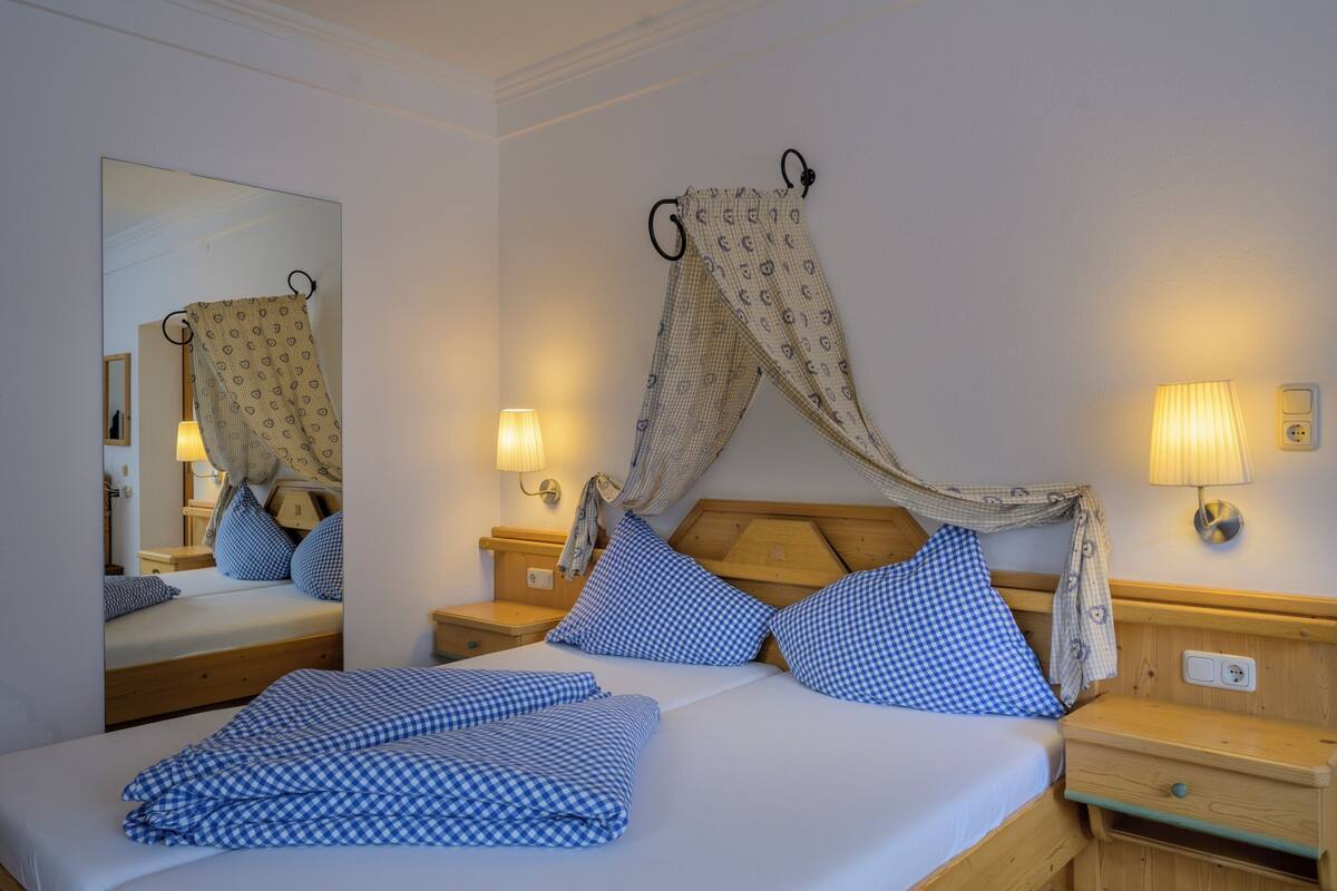 A neatly made bed with blue checkered bedding, a wooden headboard, and a canopy. There are bedside lamps and a nightstand with a lamp.