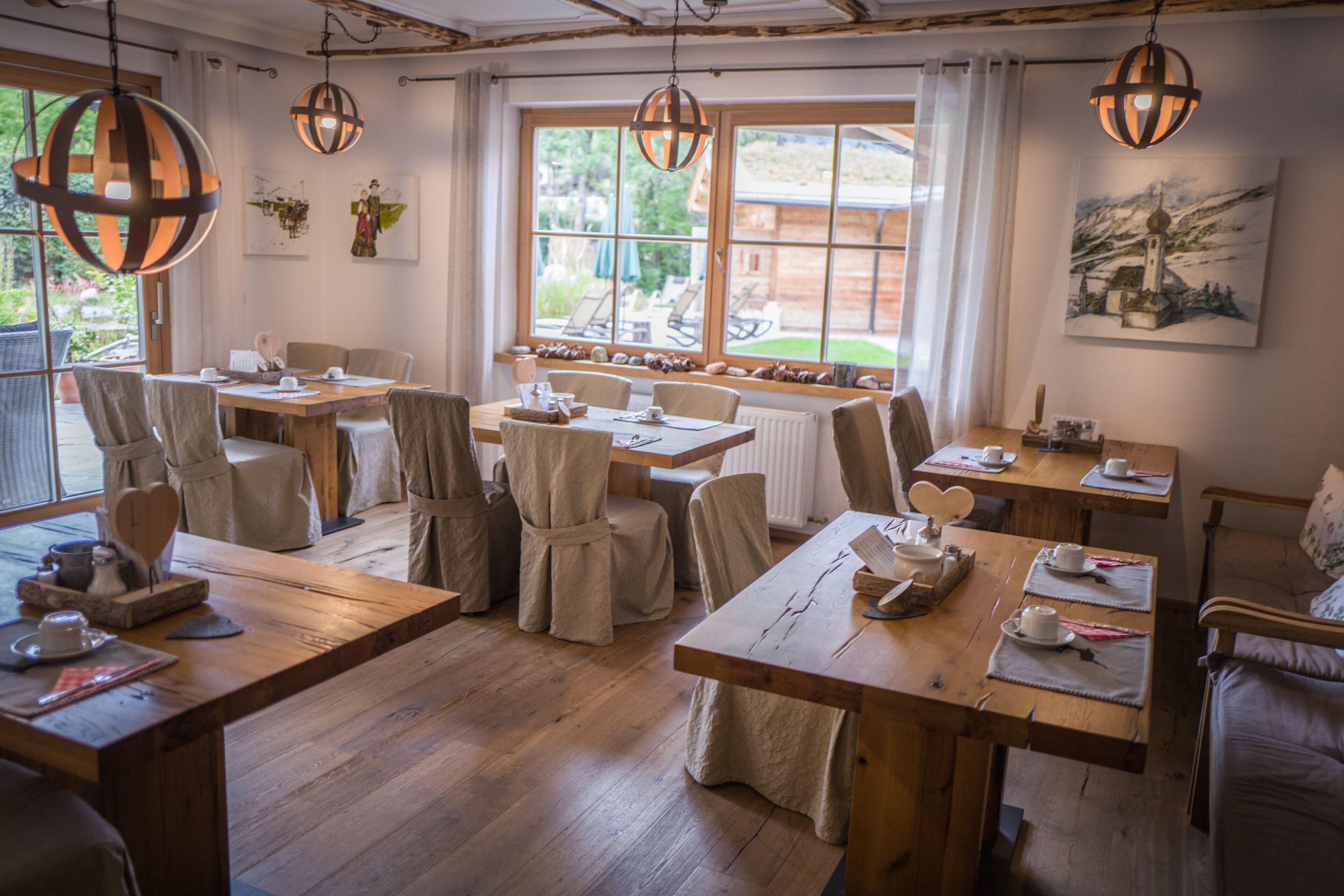 Un comedor con mesas y sillas de madera, ventanas amplias con cortinas, y lámparas esféricas colgantes.