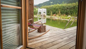 A wooden deck with a rocking chair and a small table, overlooking a pond and a garden with a white gazebo.
