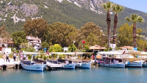 Un puerto deportivo con varias embarcaciones amarradas, un pequeño edificio de techo rojo y un paisaje montañoso al fondo.