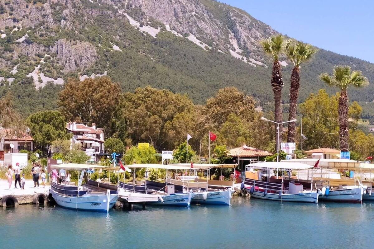 A marina with several boats docked, a small building with a red roof, and a mountainous backdrop.