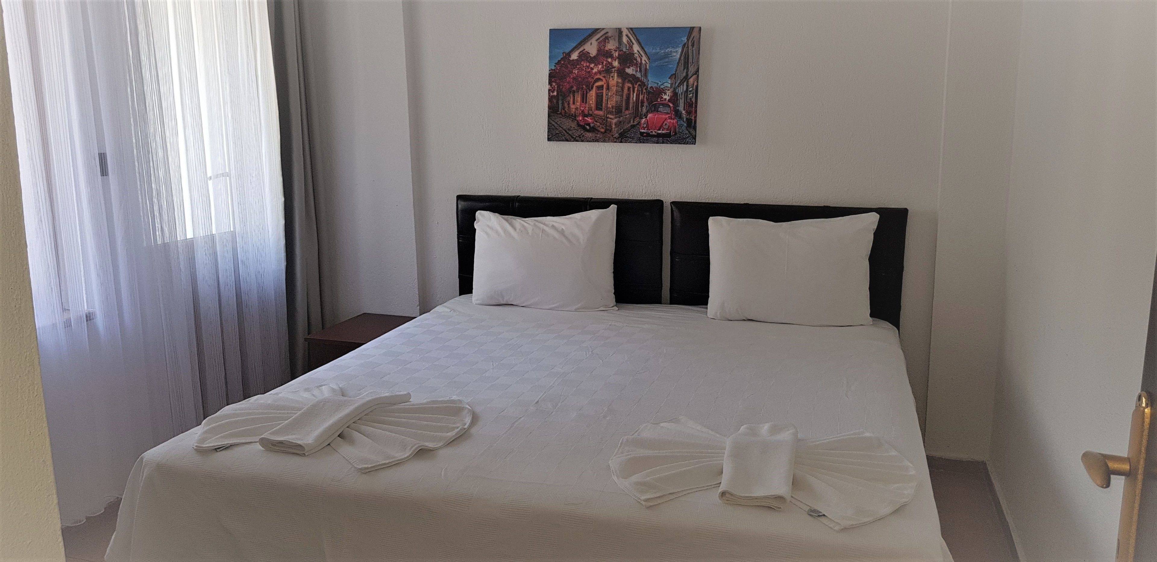 Un llit ben fet amb llençols blancs i dos coixins, un quadre a la paret i una finestra amb cortines transparents.