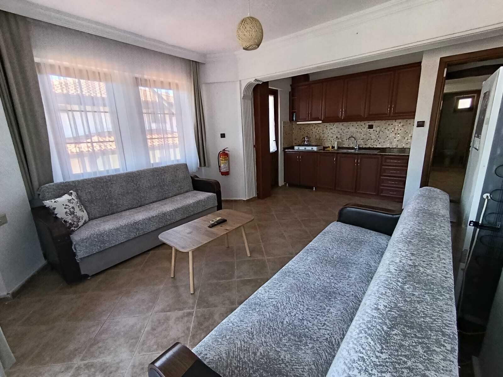 A living room with a grey sofa, a wooden coffee table, and a grey couch. There is a kitchen area with wooden cabinets and a tiled backsplash.