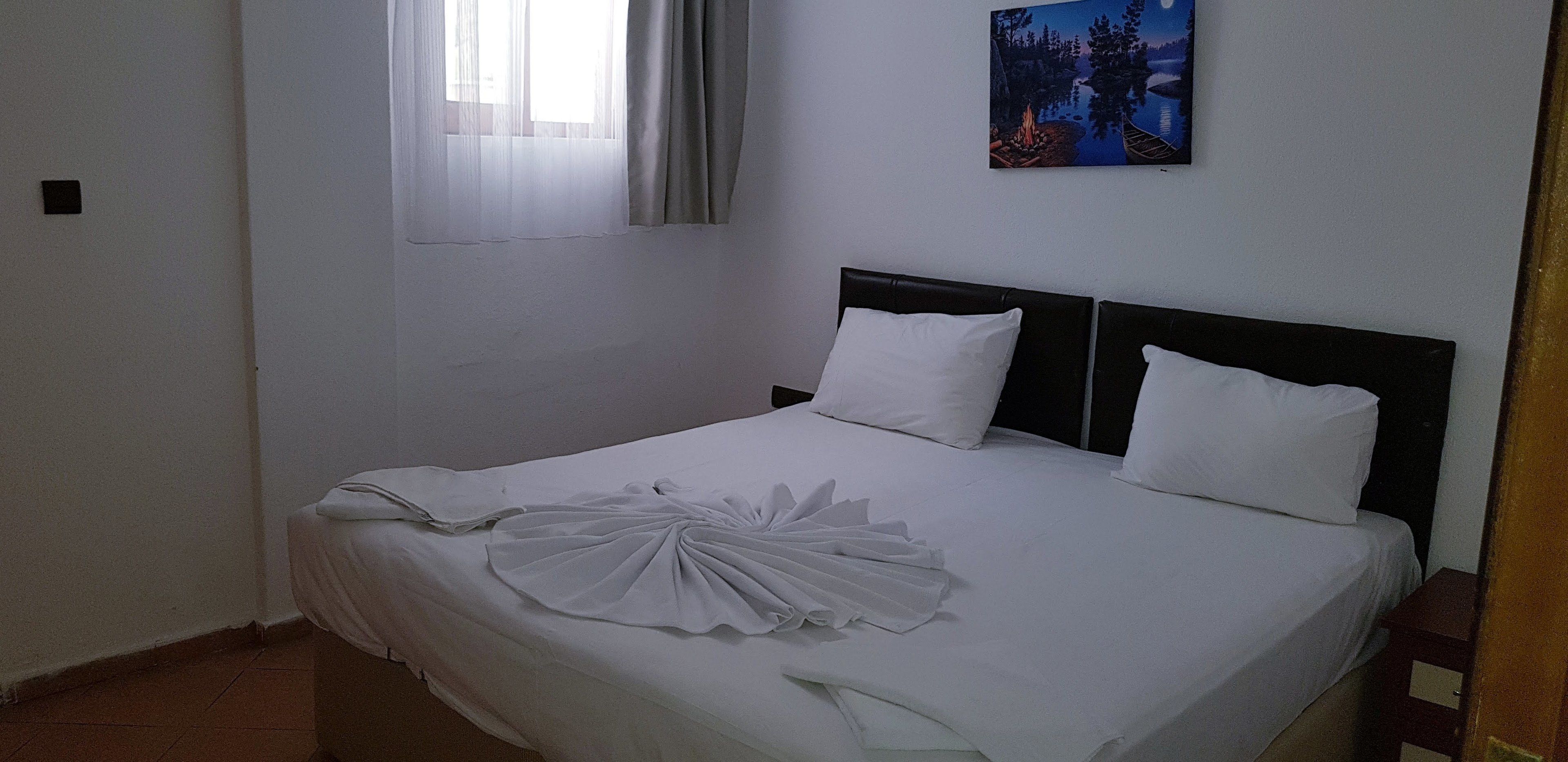 A neatly made bed with white linens and a decorative white towel. There is a painting of a forest scene on the wall above the bed.