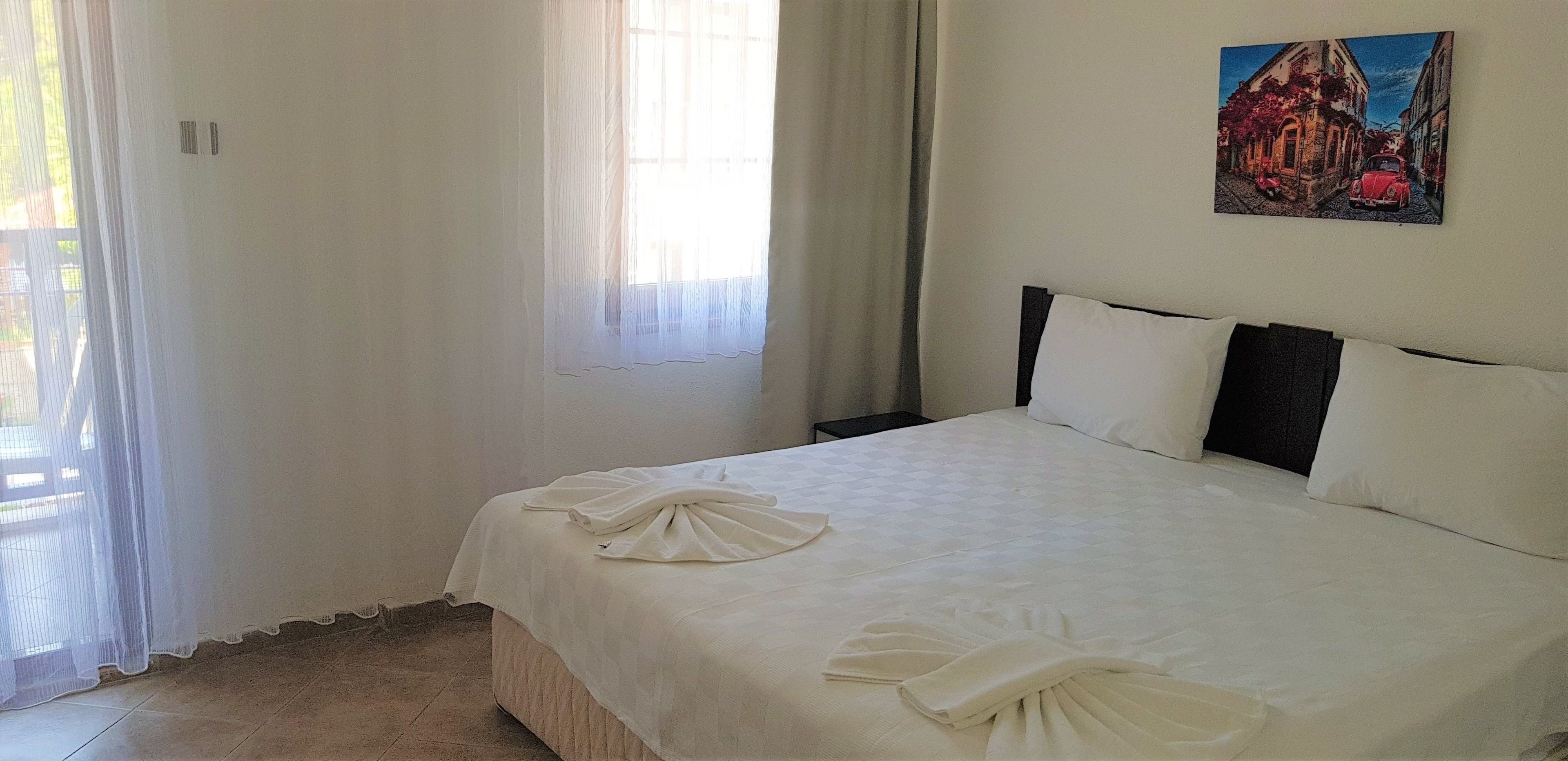 A neatly made bed with white linens and towels, a painting on the wall, and a window with sheer curtains.
