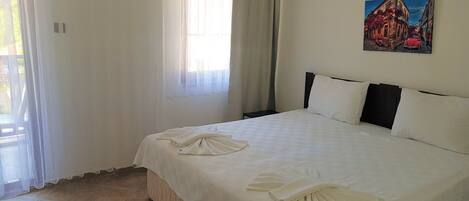 A neatly made bed with white linens and towels, a painting on the wall, and a window with sheer curtains.