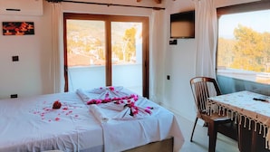 Una habitación con una cama, una mesa y una silla. Se aprecia una ventana con cortinas, un televisor montado en la pared y un marco de fotos.