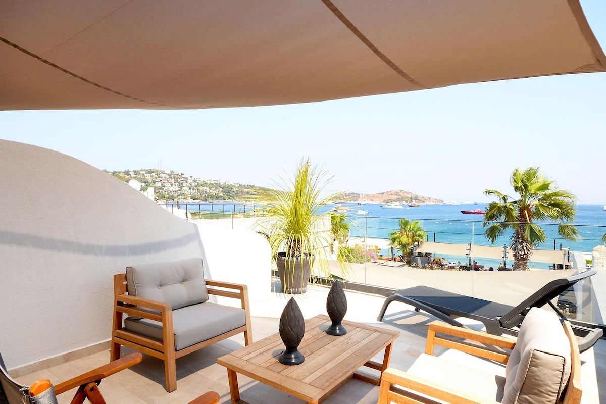 A terrace with a sofa, a table, and a lounge chair, overlooking a coastal area with a view of the sea and boats.